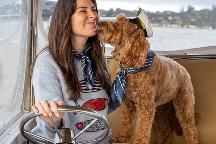 Woman with a striped scarf and dog wearing a captain's hat on a boat, as the dog licks her face.